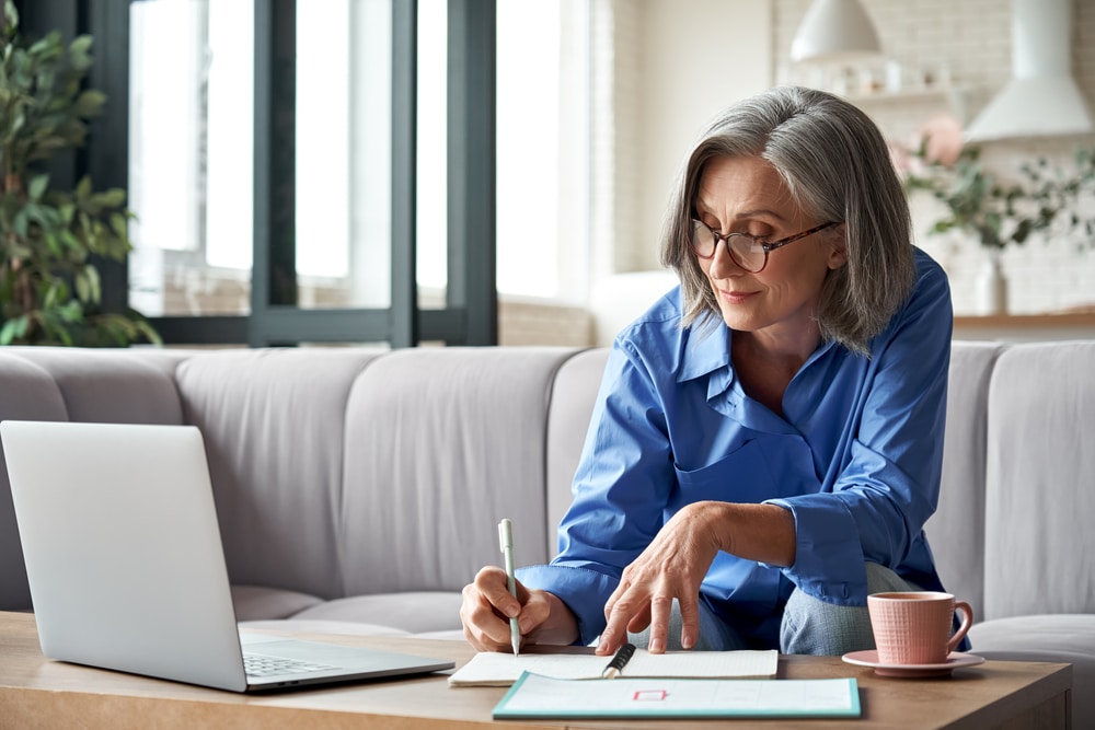 A view of an old lady writing on a memo pad with laptop on the side